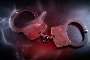 Metalik kelepçeler, dönen dumanla çevrili karanlık bir yüzeyde dinleniyor. Kanun uygulama, adalet ve kısıtlama temaları büyüleyici bir görsel anlatı ile canlandırılıyor.