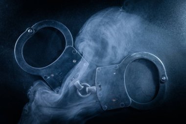 Karanlık yüzeye yerleştirilmiş kelepçeler, dumanla kaplanmış, şaşırtıcı bir görsel etki yaratıyor, adalet ve hapsedilme temalarını temsil ediyor.