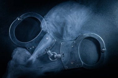 Karanlık yüzeye yerleştirilmiş kelepçeler, dumanla kaplanmış, yoğun bir ortam yaratıyor, kısıtlama, otorite ve adalet temalarını temsil ediyor.