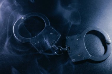 Karanlık bir yüzeye yerleştirilmiş kelepçeler, etrafta dönen dumanlar, ilgi çekici ve karamsar bir ortam yaratıyor, kısıtlama ve hapsetme temalarını temsil ediyor.