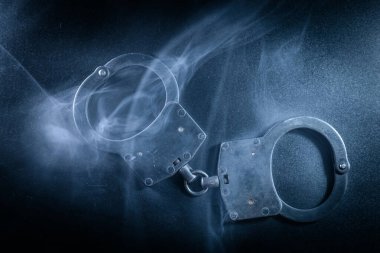 Karanlık bir yüzeye yerleştirilmiş kelepçeler, kıvrımlı dumanlarla çevrili, yasa uygulama, suç ve adaletin dengesiz bir ortamda çağrıştırdığı