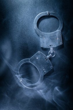 Karanlık bir yüzeye yerleştirilmiş kelepçeler, dumanla kaplanmış, otoritenin temaları ve kısıtlamaları vurgulayan gizemli bir ortam yaratıyor.