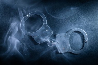 Koyu bir zemine yerleştirilmiş kelepçeler, dumanla kaplanmış, suç, adalet ve kanun uygulama temaları görsel olarak çarpıcı bir şekilde çağrıştırıyor.