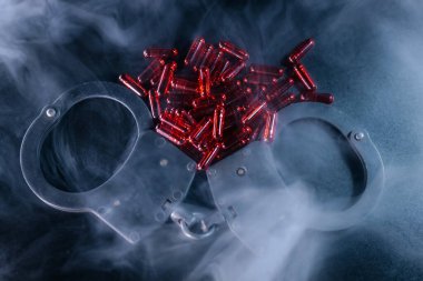 Kelepçeler karanlık yüzeyde, kırmızı kapsüller etrafa saçılmış, dumanla kaplanmış, suç, tehlike ve yasadışı maddeler çağrıştırıyor.