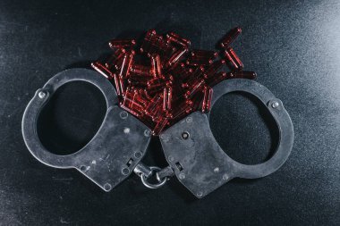 Kelepçeler kırmızı kapsül yığınıyla karanlık bir yüzeye yerleştirilir. Suç, kolluk kuvvetleri ve sonuçları temsil eden çarpıcı bir görüntü oluşturur.