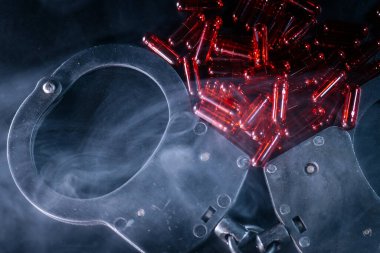 Kelepçeler, kırmızı kapsüllerin etrafa saçıldığı bir yüzeye konuluyor, dumanlı bir zemin arasında, suç, bağımlılık ve sonuçları çağrıştırıyor.