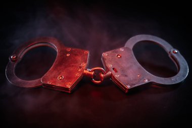 Kırmızı ışıkla aydınlatılan kelepçeler, karanlık bir zemine yerleştirilmiş, gerilim ve otorite hissi uyandırıyor, suç ve adalet temaları vurgulanıyor