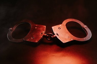 Kelepçeler koyu bir arkaplanda, sıcak bir kırmızı ışıkla vurgulanmış, kolluk kuvvetleri, adalet ve kısıtlama temaları çarpıcı bir şekilde temsil ediliyor.