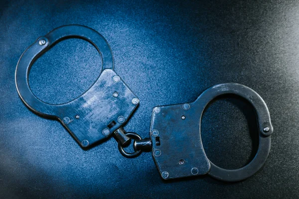 Karanlık bir yüzeye yerleştirilmiş siyah kelepçeler tasarımı ve dokusunu vurguluyor, yasa uygulama, güvenlik ve adaletin dengesiz atmosferdeki temalarını temsil ediyor.