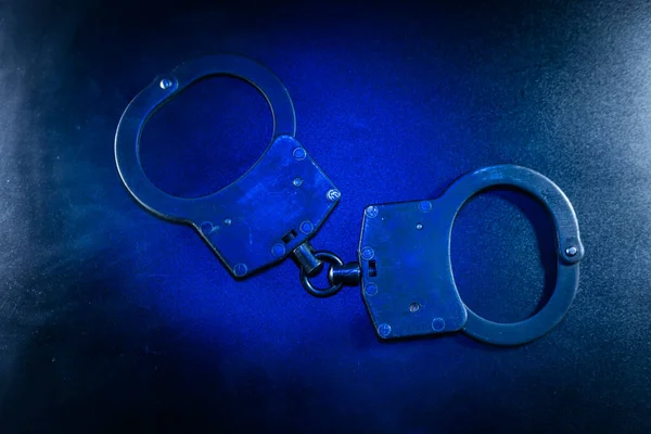 Metalik kelepçeler koyu bir yüzeyde mavi ışıkla dinleniyor, tasarımlarını ve dokusunu vurguluyor, kanun uygulama ve güvenlik temalarını temsil ediyor.