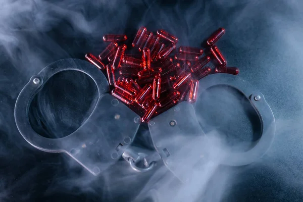 Kelepçeler karanlık yüzeyde, kırmızı kapsüller etrafa saçılmış, dumanla kaplanmış, suç, tehlike ve yasadışı maddeler çağrıştırıyor.