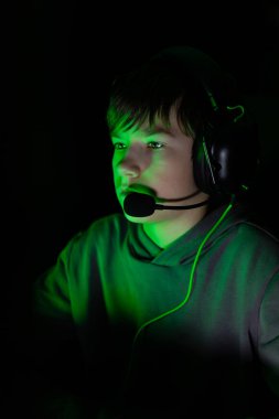 Kulaklıklı çocuk oyuna dalmış, etrafı karanlık bir çevreyle çevrili, yeşil ışıkla aydınlatılmış, yoğun bir odak ve heyecan yansıtan