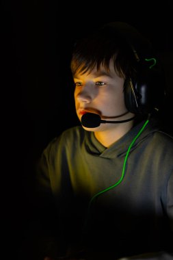 Erkek oyuncu yoğun bir şekilde karanlık bir ortamda, bir kulaklık takarak, oyun ve teknolojinin büyüleyici doğasını vurgulayarak ekrana odaklanmış durumda.