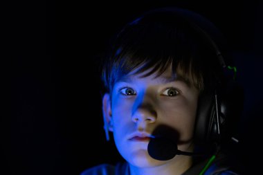 Genç oyuncu kulaklık takıyor, oyun deneyimine dalmış, etrafı karanlık ortam ve mavi ışıkla çevrili, dijital oyundaki konsantrasyon ve heyecanı ön plana çıkarıyor.