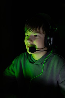 Kulaklıklı çocuk, oyun oynamaya odaklanmış, karanlık bir ortamda yeşil ışıkla aydınlatılmış, modern dijital eğlencenin ve gençlik coşkusunun özünü yakalıyor.