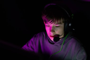 Genç erkek oyuncu bilgisayar ekranı ile meşgul, mor ışıkla aydınlatılmış karanlık bir ortamda kulaklık takıyor, oyun atmosferini vurguluyor.