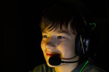 Boy wearing gaming headset is focused on screen, illuminated by soft light, creating an atmosphere of excitement and engagement in a dark environment