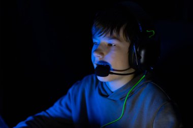 Kulaklıklı çocuk karanlık bir odada oynamaya odaklanmış, ekrandan gelen mavi ışıkla aydınlatılan, onun nişanını ve etkinliğe olan hevesini vurgulayan