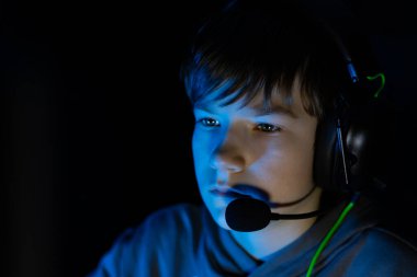 Kulaklık takan genç erkek oyuncu, yoğun bir şekilde ekrana odaklanmış, mavi ışıkla aydınlatılmış, modern oyun kültürü ve teknolojisinin özünü yakalıyor.