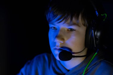 Kulaklık takan genç çocuk, derin bir şekilde oyuna odaklanmış, yumuşak mavi ışıkla aydınlatılmış, modern dijital eğlence ve büyüleyici deneyimlerin özünü yakalıyor.