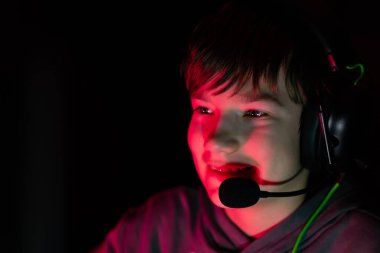 Kulaklıklı genç oyuncu ekrana odaklanıyor, kırmızı ışıkla aydınlatılıyor, dijital eğlencenin özünü yakalıyor ve karanlık bir ortamda derin deneyimler yaşıyor.