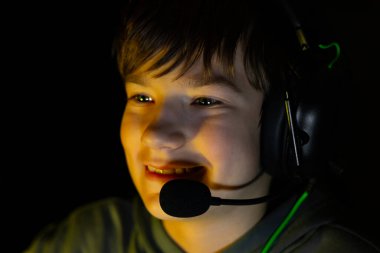 Boy wearing headset is focused on gaming, illuminated by screen light in a dark environment, capturing the essence of digital entertainment and youthful enthusiasm