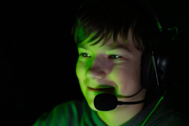 Boy wearing headphones and microphone, illuminated by green light, is focused on gaming, displaying enthusiasm and engagement in a dimly lit setting