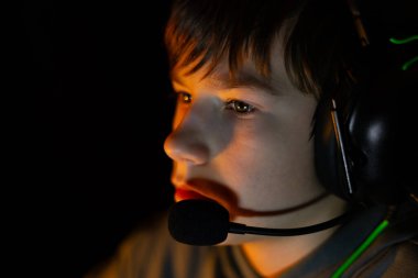 Oyun kulaklığı takan çocuk ekrana derinden odaklanmış, yumuşak ışıklandırma, dijital oyunların heyecanını ön plana çıkaran bir atmosfer yaratıyor.