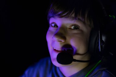 Happy young boy wearing headset, smiling in a dimly lit environment, surrounded by vibrant colors, reflecting enthusiasm for gaming and digital experiences