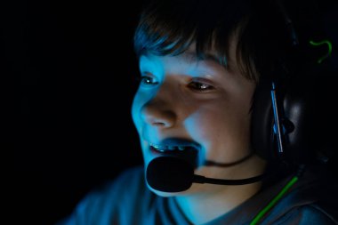 Boy wearing gaming headset is focused on screen, illuminated by blue light, creating an immersive atmosphere, reflecting the excitement of online gaming experiences