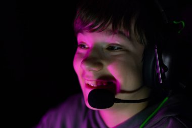 Boy wearing gaming headset, illuminated by vibrant lights, is focused on screen while playing video games, capturing the essence of youth and digital culture