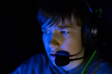 Oyun kulaklığı takan çocuk yoğun bir şekilde ekrana odaklanmış, mavi ışıkla aydınlatılmış, oyun ve dijital etkileşim için derin bir atmosfer yaratıyor.