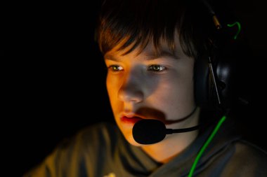 Kulaklıklı çocuk yoğun bir şekilde loş bir ortamda oyuna odaklanmış, ekran ışığıyla aydınlatılmış, dijital eğlenceye olan ilgisini ve tutkusunu vurguluyor.