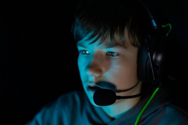 Kulaklık takan çocuk yoğun bir şekilde oyuna odaklanmış, yumuşak mavi ışıkla aydınlatılıyor, modern teknolojiyi ve karanlık ortamda deneyimi ön plana çıkarıyor.