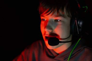 Kulaklıklı erkek oyuncu, kırmızı ışıkla aydınlatılmış, ekrana odaklanmış, rekabetçi oyunların özünü yakalıyor ve dijital eğlencenin heyecanını yakalıyor