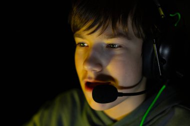 Kulaklık takan çocuk oyuna odaklanmış, karanlık bir ortamda ekran ışığıyla aydınlatılıyor, dijital etkileşimin ve gençlik coşkusunun özünü yakalıyor.