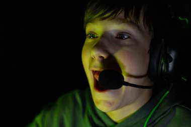 Excited young gamer wearing headset is immersed in video game experience, surrounded by dark atmosphere and vibrant green lighting, capturing the joy of gaming