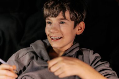 Gri kapüşonlu çocuk gülümsüyor ve elinde bir kalem tutuyor. Yumuşak ışıklandırmalı rahat bir ortamda neşe ve yaratıcılık anı yakalıyor.