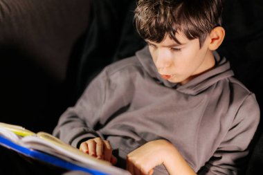 Gri kapüşonlu genç bir çocuk kanepede otururken kitap okumakla meşgul, rahat bir atmosferi ve öğrenme sevincini ön plana çıkarıyor.