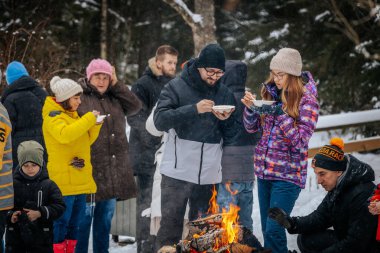 Ligatne, Letonya - 11 Ocak 2026: Karlı bir ormanda kamp ateşi altında çorbanın tadını çıkaran çeşitli kişiler grubu, sıcak bir kış ortamı yaratıyor.