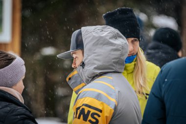 Ligatne, Letonya - 11 Ocak 2026: Gri ve sarı ceketli bir adam karlı açık havada arkadaşlarıyla takılıyor, kış kıyafetlerini ve arkadaşlığını ön plana çıkarıyor