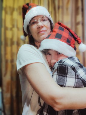 Kırmızı ve siyah ekose Noel Baba şapkalı kadın ve çocuk, neşeli bir bayram anında, yumuşak dokular ve sıcak aydınlatmalarla çevrili evde kucaklaşıyorlar.
