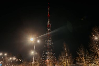 Haberleşme kulesi gece boyunca ayakta duruyor, kırmızı ve beyaz ışıklarla aydınlatılıyor, sokak lambaları ve ağaçlarla çevrili.