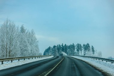 Karla kaplı ağaçlarla çevrili dolambaçlı bir yol ve uzak bir ormanda bulutlu bir gökyüzüne karşı sakin bir kış manzarası.