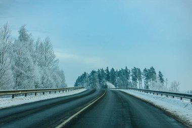 Karla kaplı ağaçlarla çevrili dolambaçlı yol bulutlu bir gökyüzünün altında uzak bir ormana çıkar ve sakin bir kış manzarası yaratır.