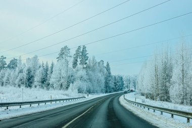 Karla kaplı ağaçlar ve elektrik hatlarıyla kaplı dolambaçlı yol soluk mavi gökyüzünün altında sakin bir kış sahnesi yaratıyor.