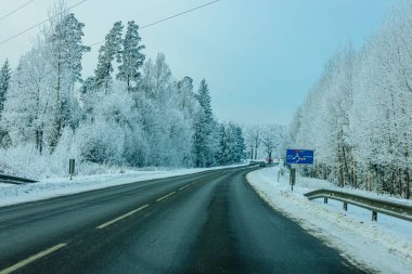 Valmiera, Letonya - 21 Ocak 2026: Kenarlarında buzlu ağaçlar ve mavi bir işaret olan kış manzarası boyunca karlı yol kıvrımları