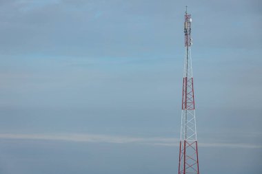 Kırmızı ve beyaz çizgili iletişim kulesi bulutlu mavi gökyüzüne yükseliyor, açık alanda modern telekomünikasyon altyapısını temsil ediyor.
