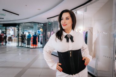 Beyaz bluzlu ve siyah korseli bir kadın bir alışveriş merkezinde duruyor, arka planda moda eşyaları ve kıyafet sergileri sergiliyor.