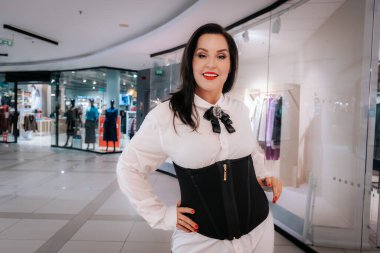 Beyaz kadın model, beyaz bir bluzun üzerinde siyah bir korse sergiliyor. Çağdaş bir alışveriş merkezinin içinde. Arka planda giysi ekranları görünüyor.